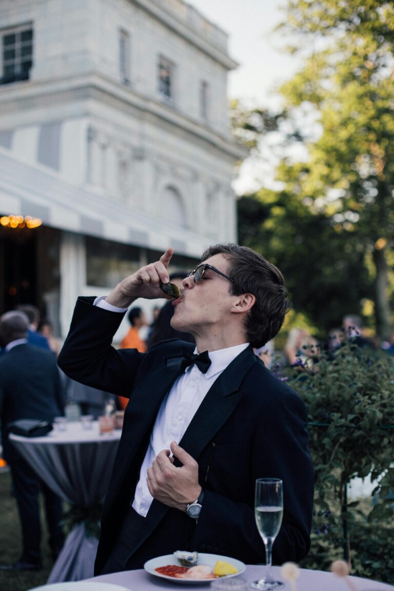 Sophisticated man in black suit enjoys oysters at a lively outdoor gathering in Newport.